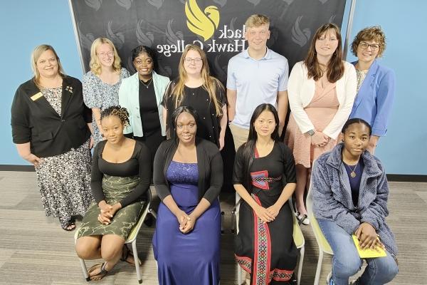 7 people standing and 4 people sitting in front of Black Hawk College logo backdrop