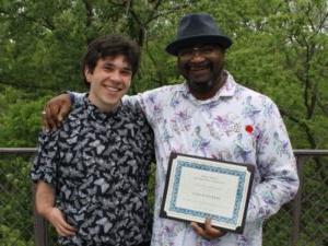 2 people standing outside with trees in background and one person holding a certificate