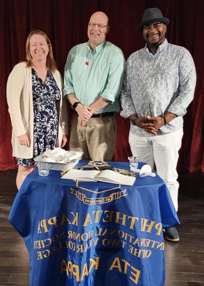 3 people standing on stage in front of small draped with Phi Theta Kappa banner